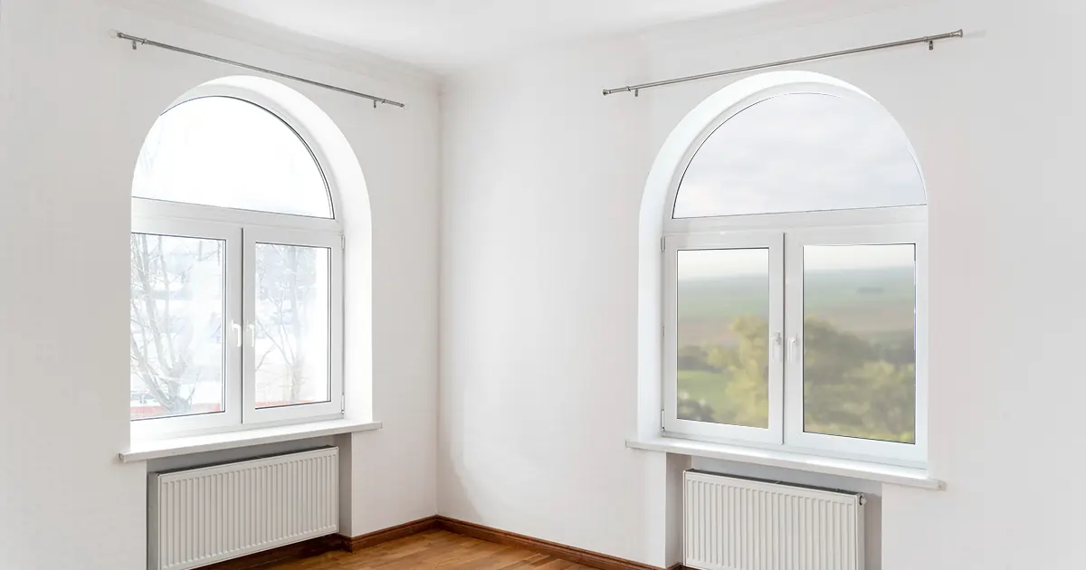 A bright, empty room with two large windows with different views, a wooden floor, and white walls.